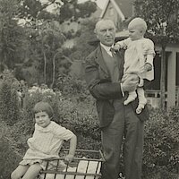 Zwei Kleinkinder und ein Mann, in einem Garten vor einem Haus und Gartenstühlen