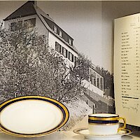 Vitrine mit Fotografie eines Hauses, einem feinen weißblauen Teeservice und einer Gästeliste