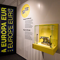 Ausstellungswand mit Vitrine, in der sich eine Bronzestatue der römischen Wölfin befindet; in gelber Schrift ist im Hintergrund "Europa" zu lesen 