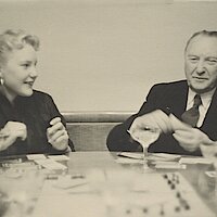 Libet Werhahn und ihr Vater Konrad Adenauer beim Monopoly-Spiel. Auf dem Tisch stehen Gläser und das Spielbrett, Libet (links) wendet sich ihrem Vater zu. Adenauer (rechts) hält Spielgeld in der Hand.
