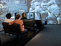 Drei Personen sitzen in einem nachgebauten Lincoln. An großen Bildschirmen sieht man eine Menschenmenge und Konrad Adenauer.