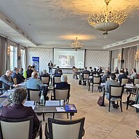 gro&szlig;er Sitzungssaal mit Blick auf Rednerpult und Leinwand, zahlreiche Teilnehmer auf St&uuml;hlen