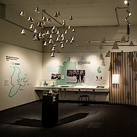 Ausstellung mit Tischen, Objekten, Fotos und Freiheitsglocken, die von der Decke hängen