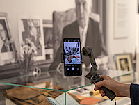 eine Hand hält ein Smartphone an einem Gimbal, im Hintergrund sieht man Fotos von Adenauer und Ausstellungsobjekte