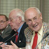 Drei Männer sitzen am Konferenztisch, ein Mann lächelt 