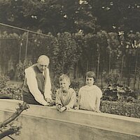 Ein Mann auf einem Mäuerchen sitzend, neben ihm stehen zwei Kleinkinder