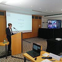 Eine Frau spricht am Rednerpult. Im Hintergrund an einem Bildschirm werden Gäste einer Videokonferenz angezeigt.