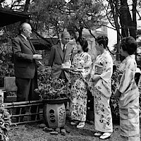 Konrad Adenauer im Garten eines Gästehauses in Japan, gemeinsam mit einer Gruppe Geishas und einem jungen Mann.