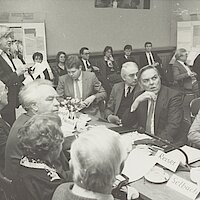 Männer und Frauen sitzen an Konferenztischen, eine Frau spricht in ein Mikrofon